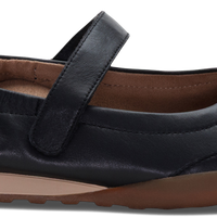 A black leather Mary Jane flat shoe with a hook-and-loop strap, brown insole, and brown outsole, shown from a side angle on a white background.